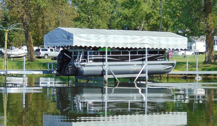 Pontoon boat on a covered lift at a lake dock, surrounded by trees and parked cars in the background.