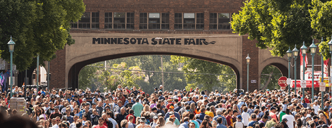 Crowd at Minnesota State Fair entrance, bustling with visitors under clear skies, highlighting popular summer event.
