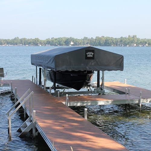 Covered boat docked at a wooden pier on a calm lake with a tree-lined shore in the background.
