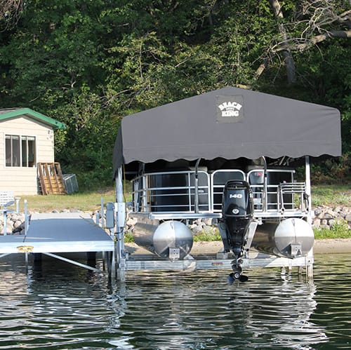 Pontoon boat on lift at lakeside, covered with canopy, next to dock, with trees and cabin in the background.