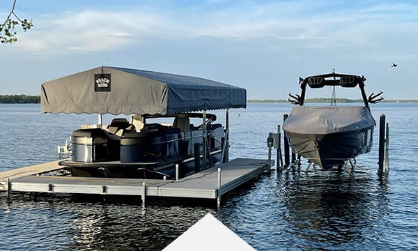 Covered pontoon boat and wakeboard boat docked on a calm lake, sunny weather.
