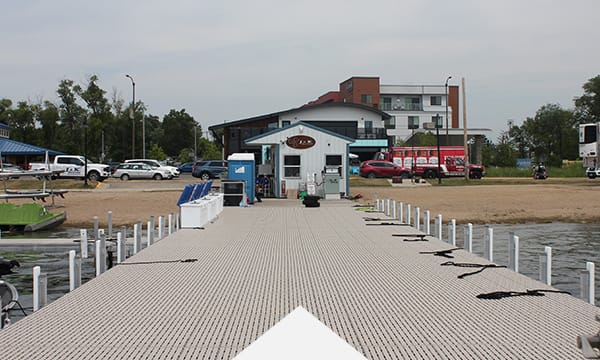 Boat dock leading to a small building near a beach, surrounded by parked cars and trees, with apartments in the background.