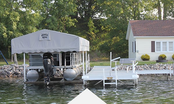 Pontoon boat docked by a lakeside house with a covered lift under trees.